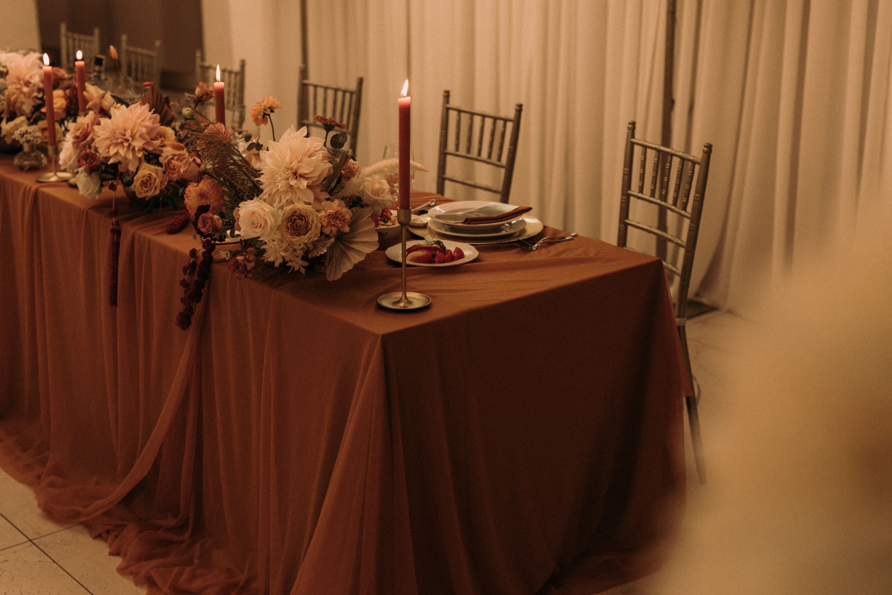 Stół pary młodej z kolorowym obrusem oraz falbaną. Na stole niskie dekoracje z szarłatem, daliami oraz różami