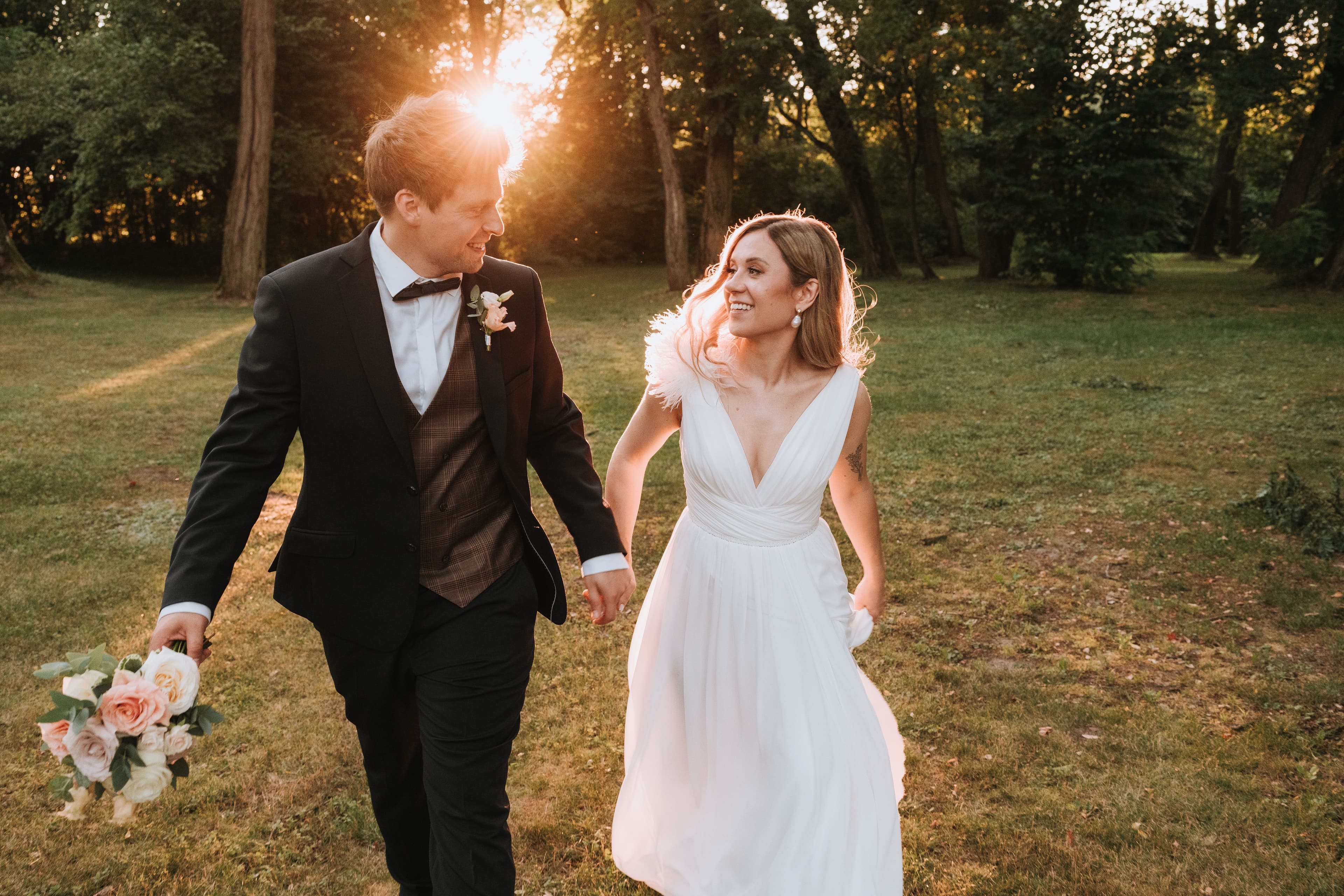 Para Młoda spacerująca po trawniku, mężczyzna w czarnym garniturze z butonierką, kobieta w białej sukni ślubnej trzymająca bukiet kwiatów w jasnej palecie