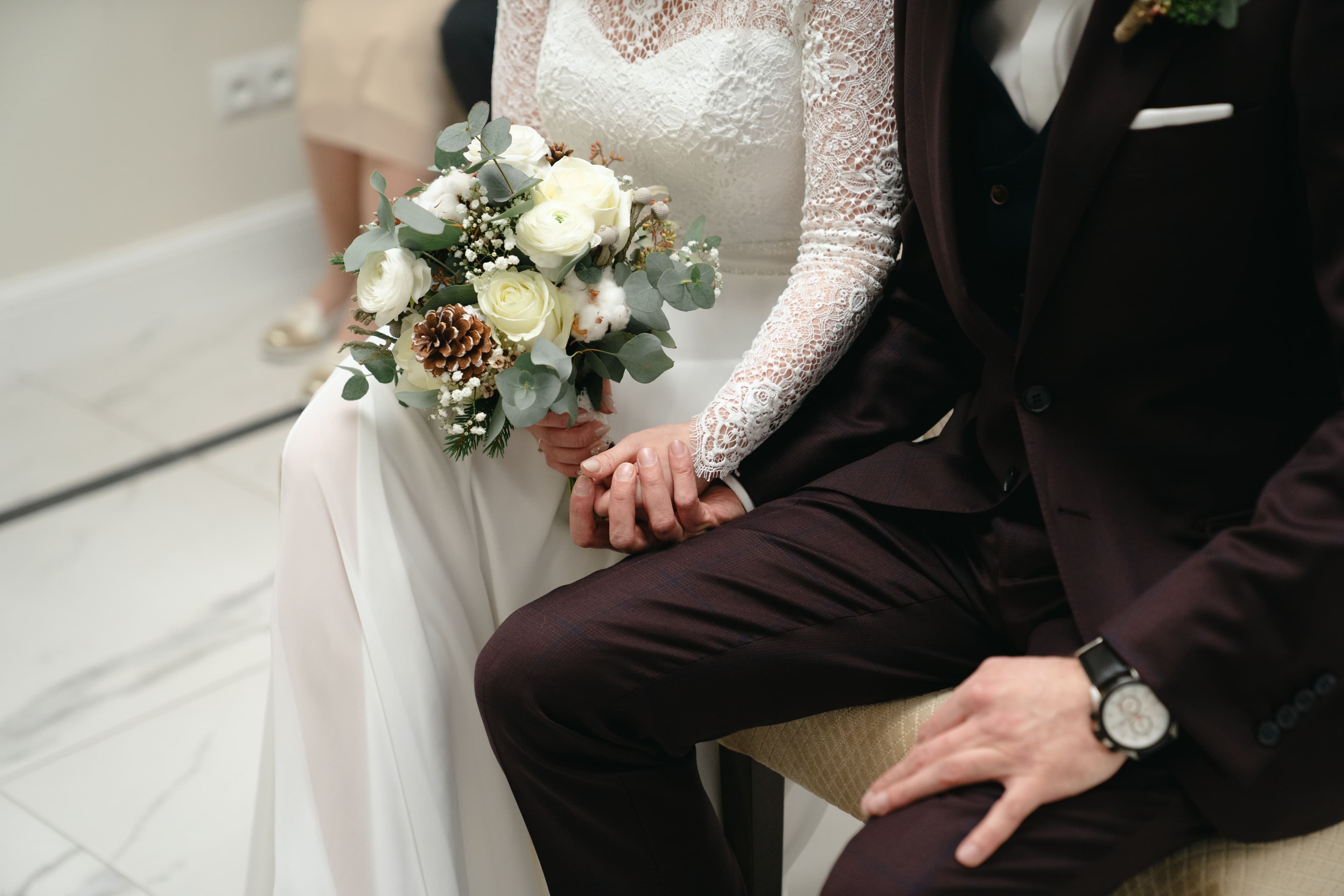 Bukiet ślubny z białymi kwiatami, zielenią i szyszkami, trzymany przez pannę młodą w koronkowej sukni i pana młodego w ciemnym garniturze.