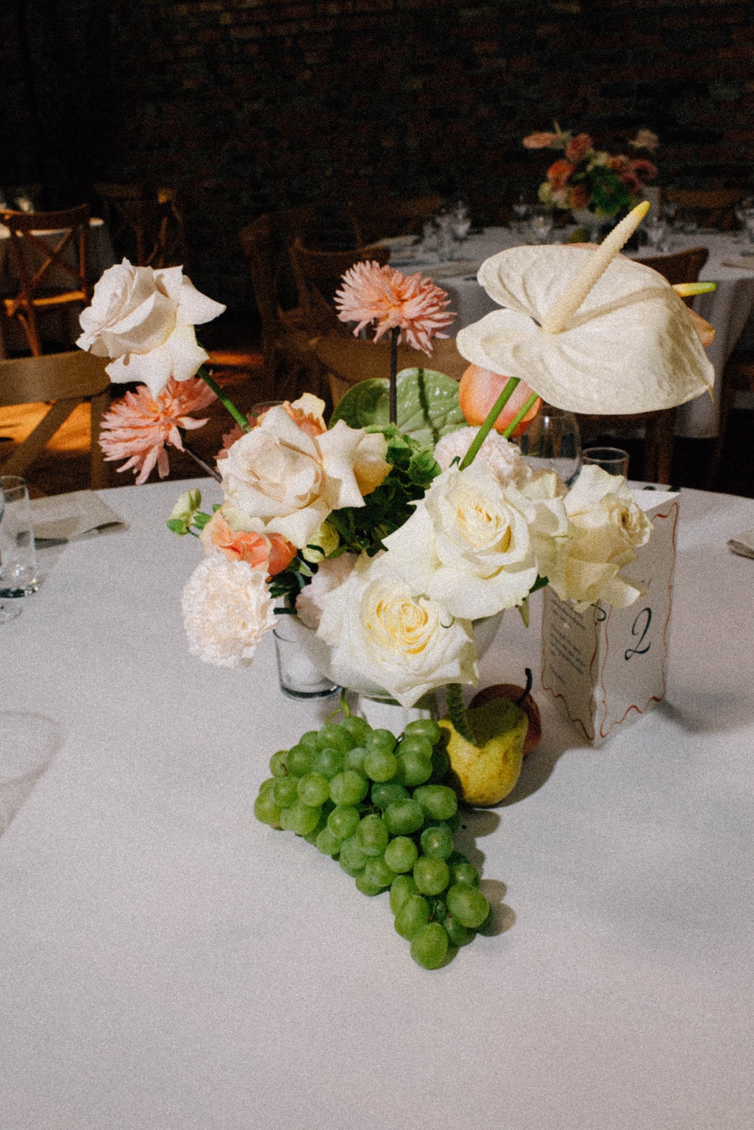 Kompozycja kwiatowa na stół z białymi różami, anthurium, pastelowymi gerberami, zielenią i owocami na białym obrusie