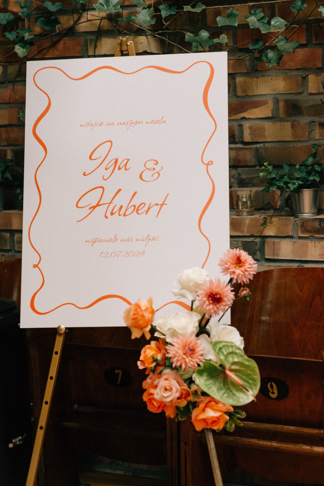 Papeteria ślubna z pomarańczową ramką, imionami Iga & Hubert, datą 12.07.2024 na tle ceglanego muru z bluszczem i bukietem kwiatów w pomarańczowych i białych tonach.