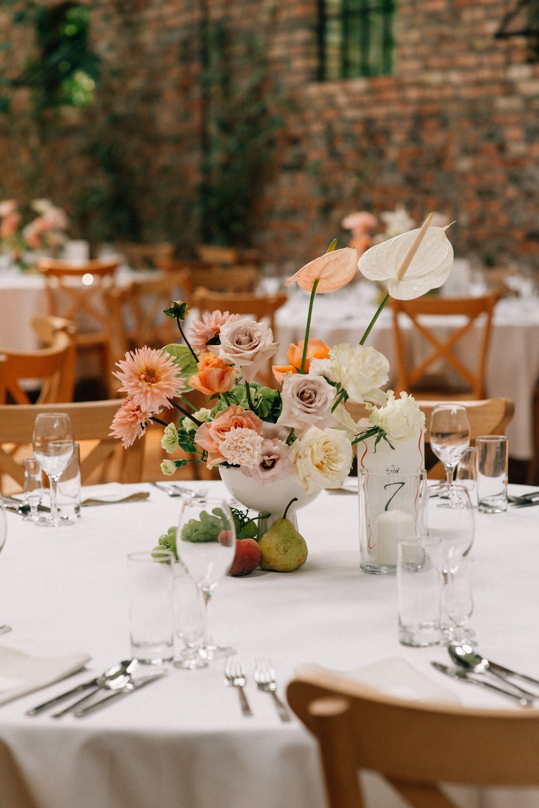 Kompozycja kwiatowa na stół z różami, gerberami i anturiami w pastelowych tonach, owoce, numerk stołu, drewniane krzesła, ceglana sala