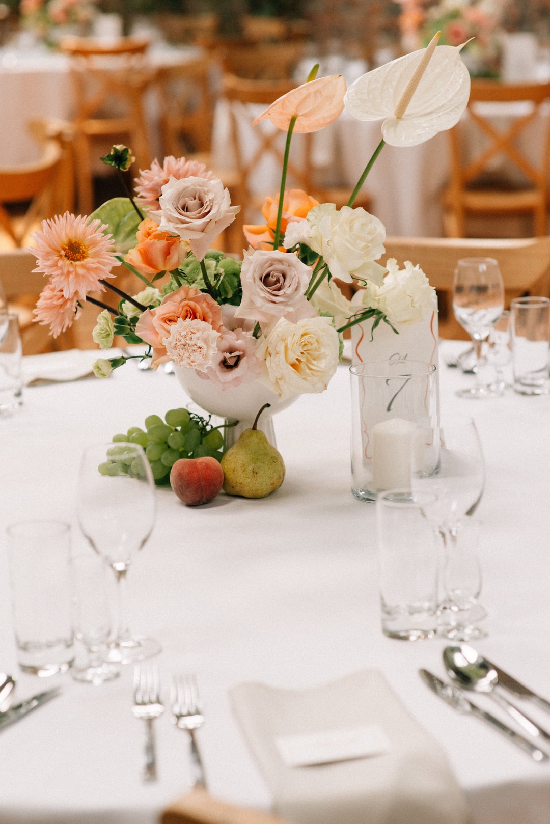 Kompozycja kwiatowa na stół w białej wazie z różami, gerberami i anturiami w pastelowych tonach, z owocami przy podstawie, na białym obrusie.