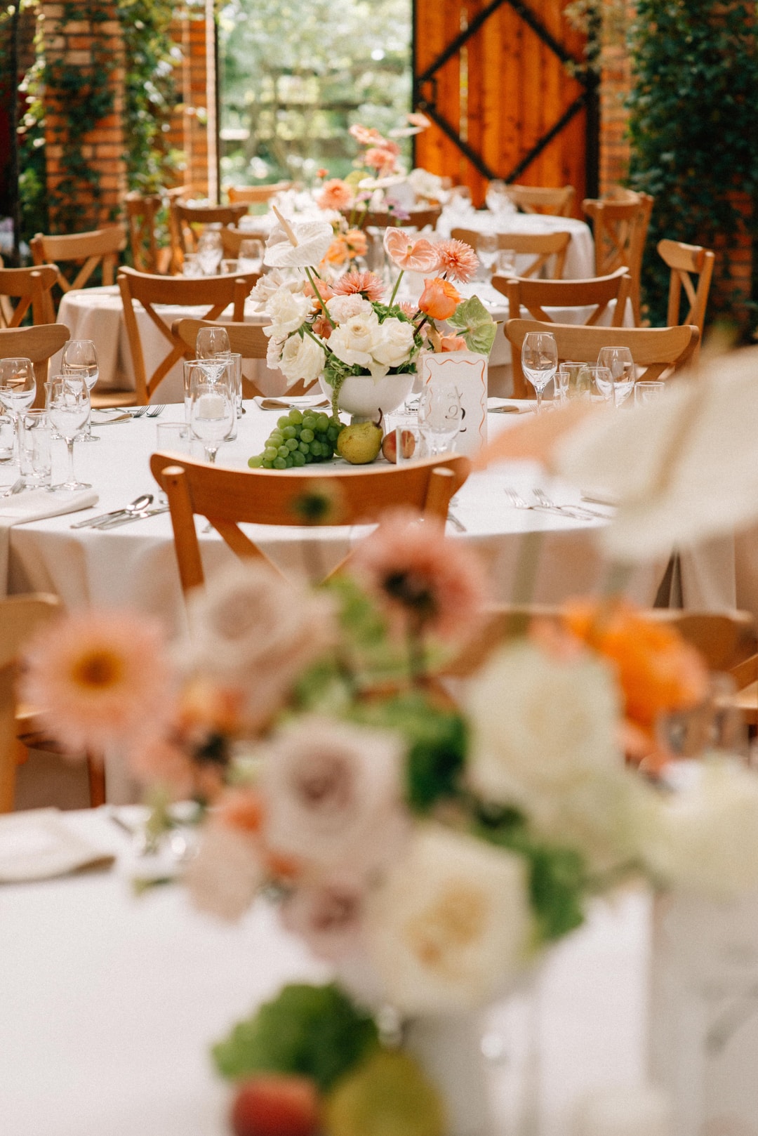 Aranżacja stołów weselnych z białymi obrusami, drewnianymi krzesłami, kompozycjami kwiatowymi w jasnej palecie i zielenią, kieliszkami i sztućcami.