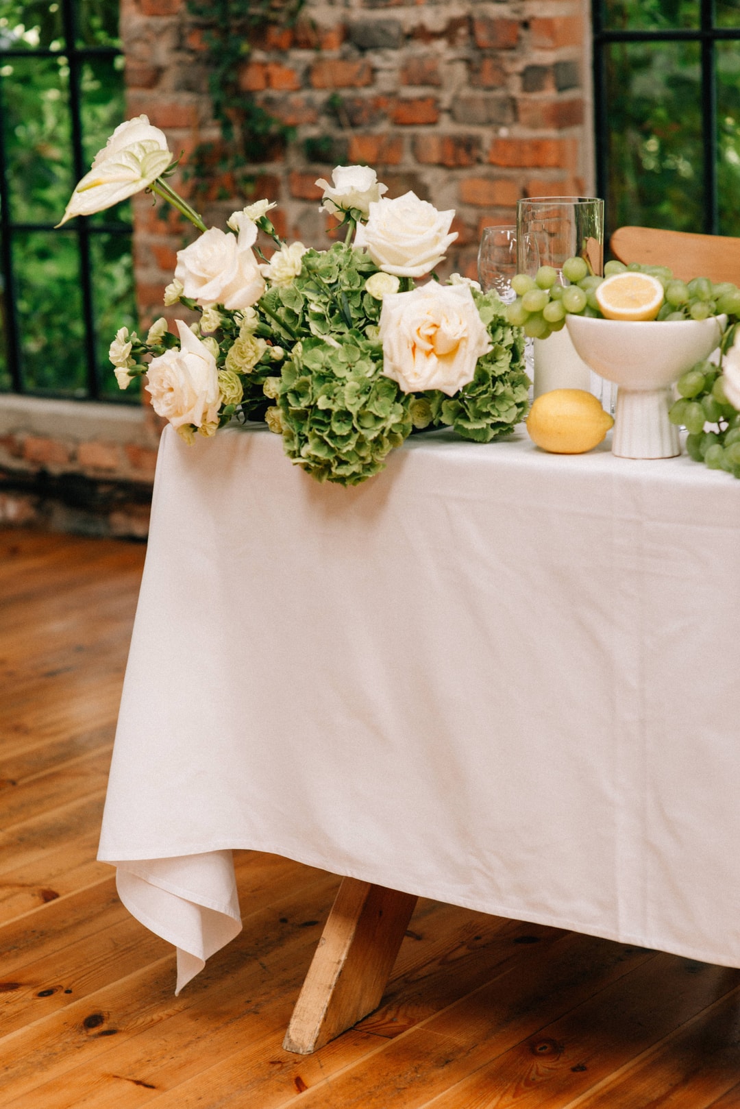 Kompozycja kwiatowa na stół z białymi różami, zielenią hortensji, szklankami wody i owocami na lnianym obrusie
