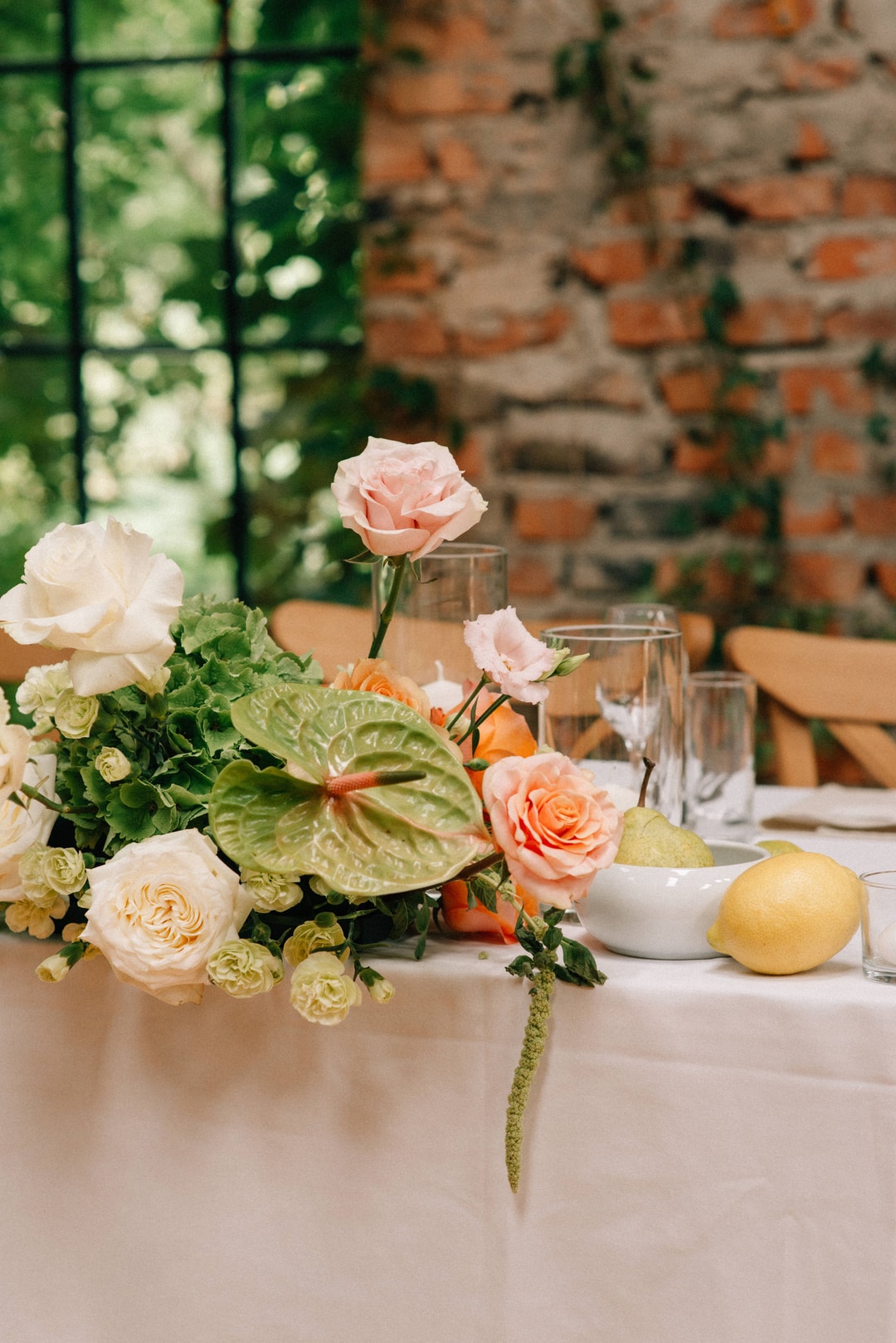 Kompozycja kwiatowa na stół z różowymi i białymi kwiatami, zielonymi liśćmi, na tle ceglanej ściany i zielonych drzwi