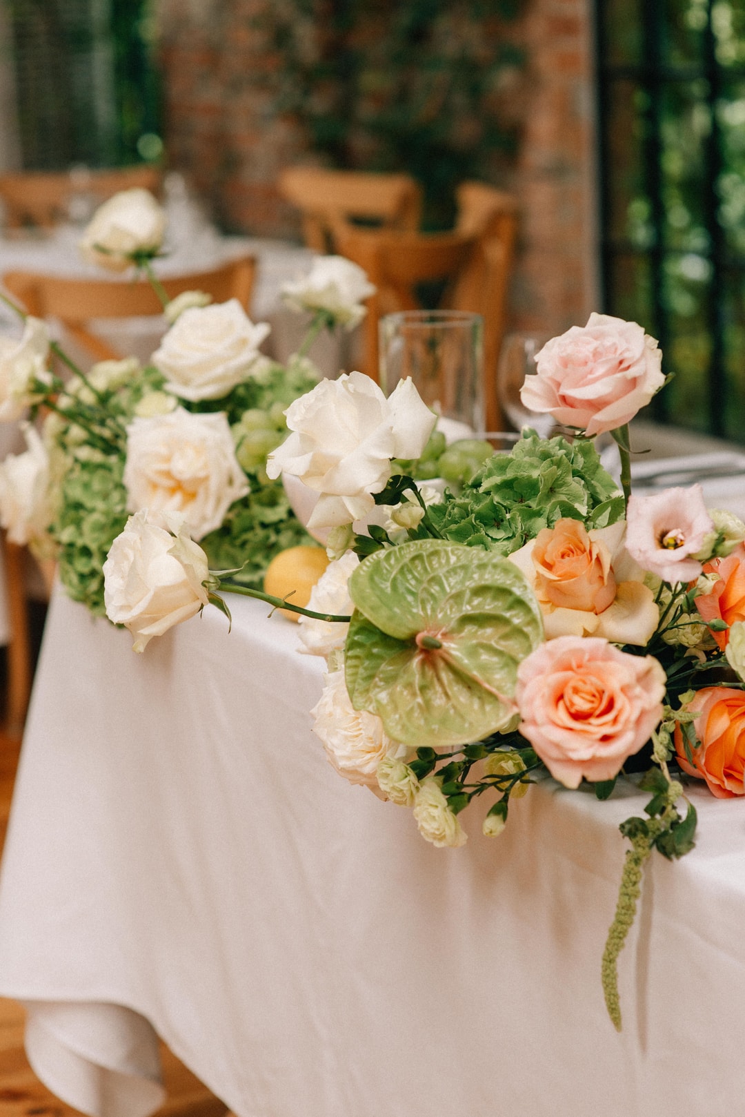 Kompozycja kwiatowa na stół z białymi różami, różowymi kwiatami, zielenią i anturium na białym obrusie, drewniana sala w tle