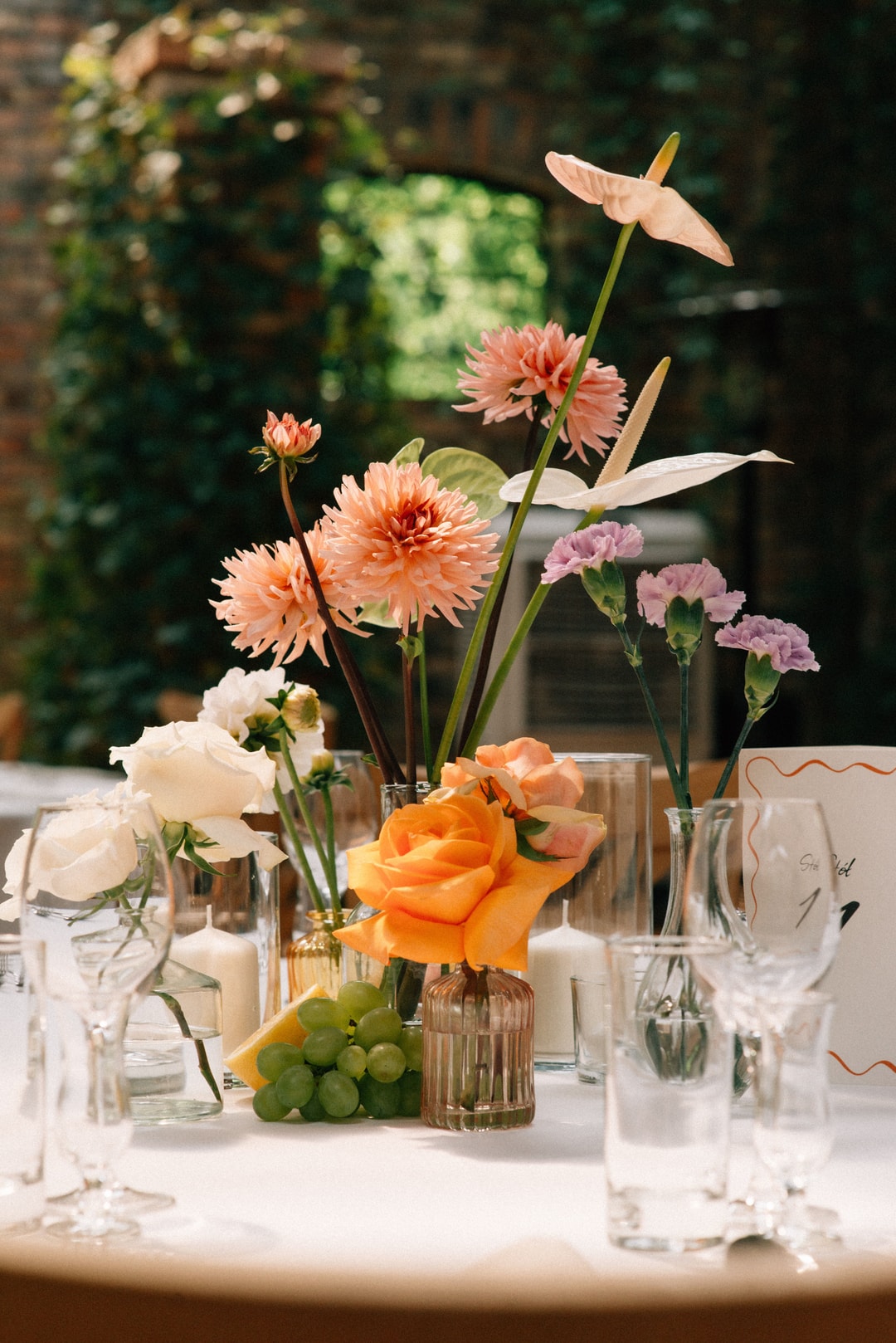 Kompozycja kwiatowa na stół z łososiowymi dalią, pomarańczową różą, różowymi goździkami i liśćmi w szklanych wazonach, na tle zieleni