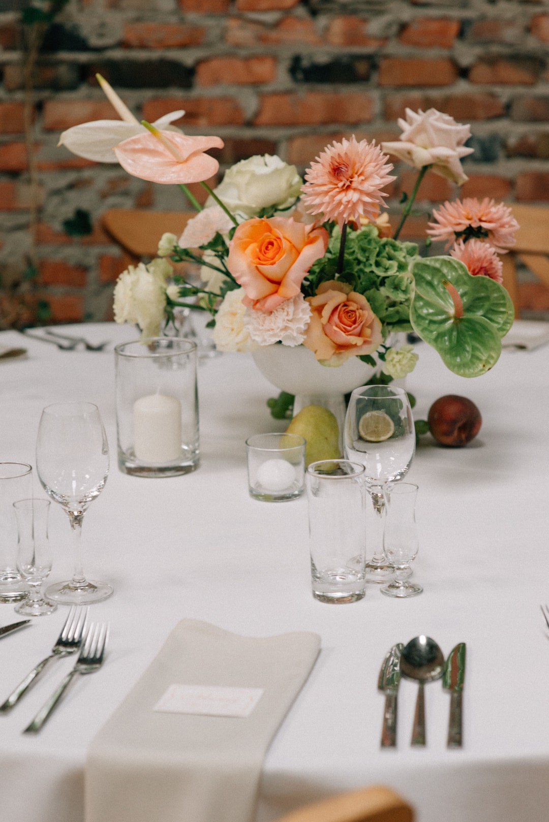 Kompozycja kwiatowa na stół z różami w tonach pomarańczowych, białymi kwiatami, gerbera i zielenią, na białym obrusie z sztućcami i szklankami, na tle ceglanej ściany