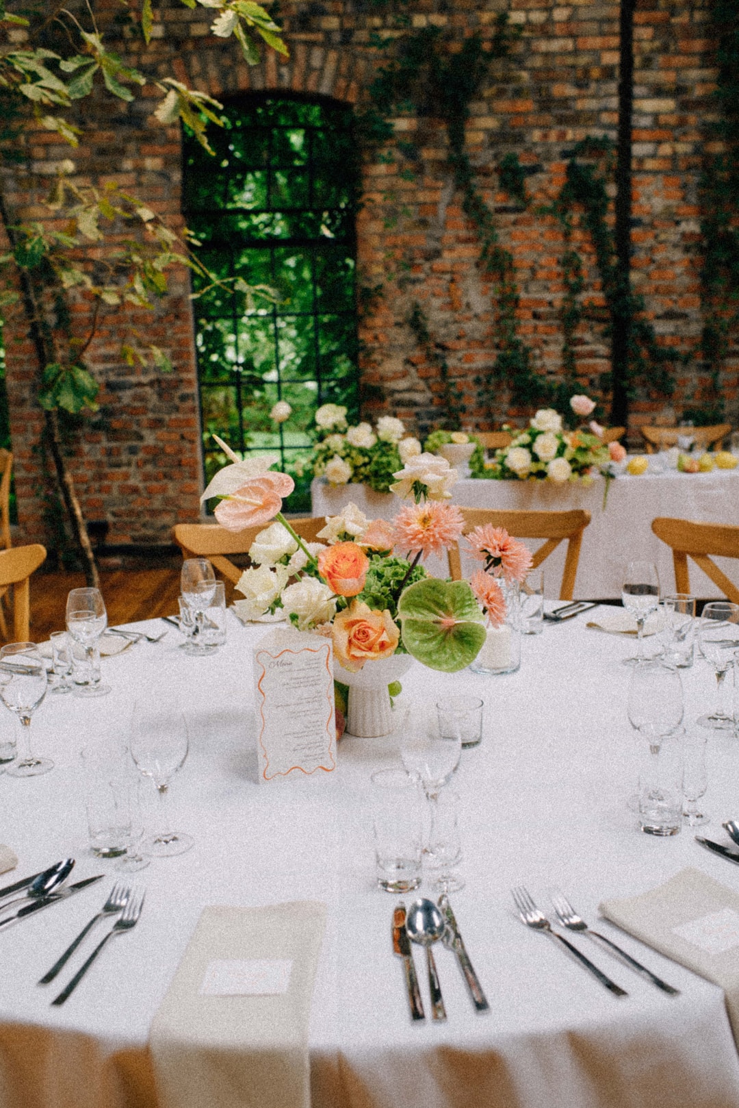 Dekoracja stołu weselnego z kompozycją kwiatową w jasnych tonach, drewnianymi krzesłami i białym obrusem, na tle ceglanej ściany z zielenią.