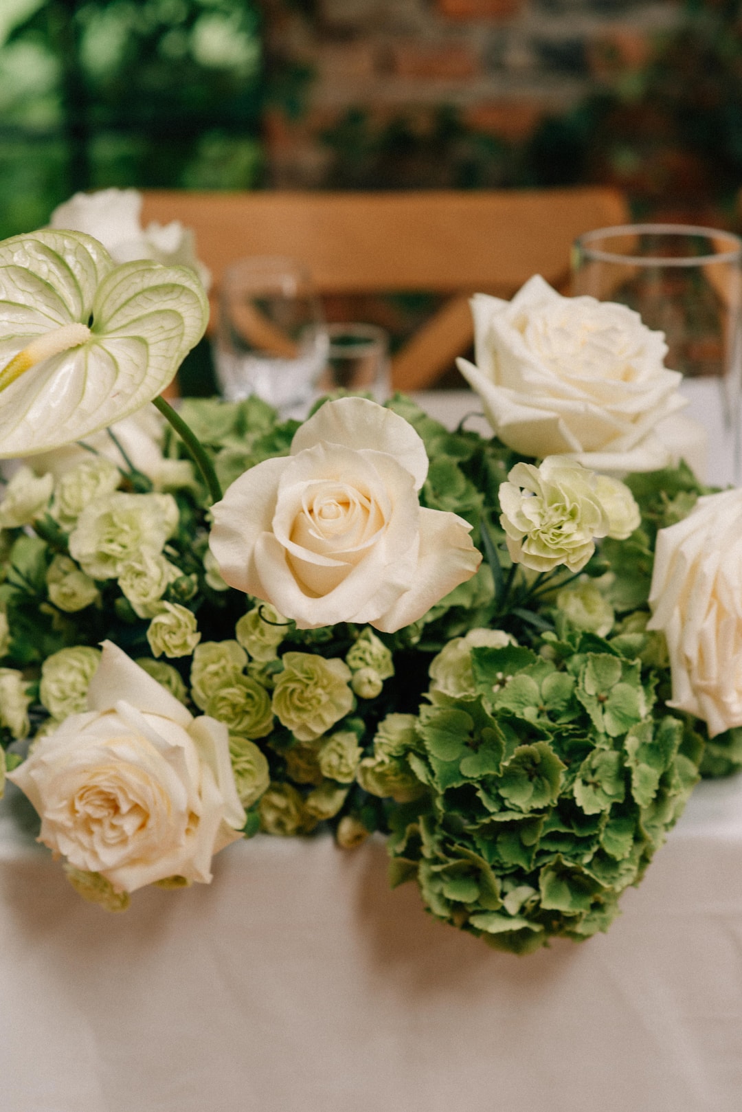 Kompozycja kwiatowa na stół z kremowymi różami, zielenią hortensji i białymi liśćmi, na jasnym tle drewnianej sali.