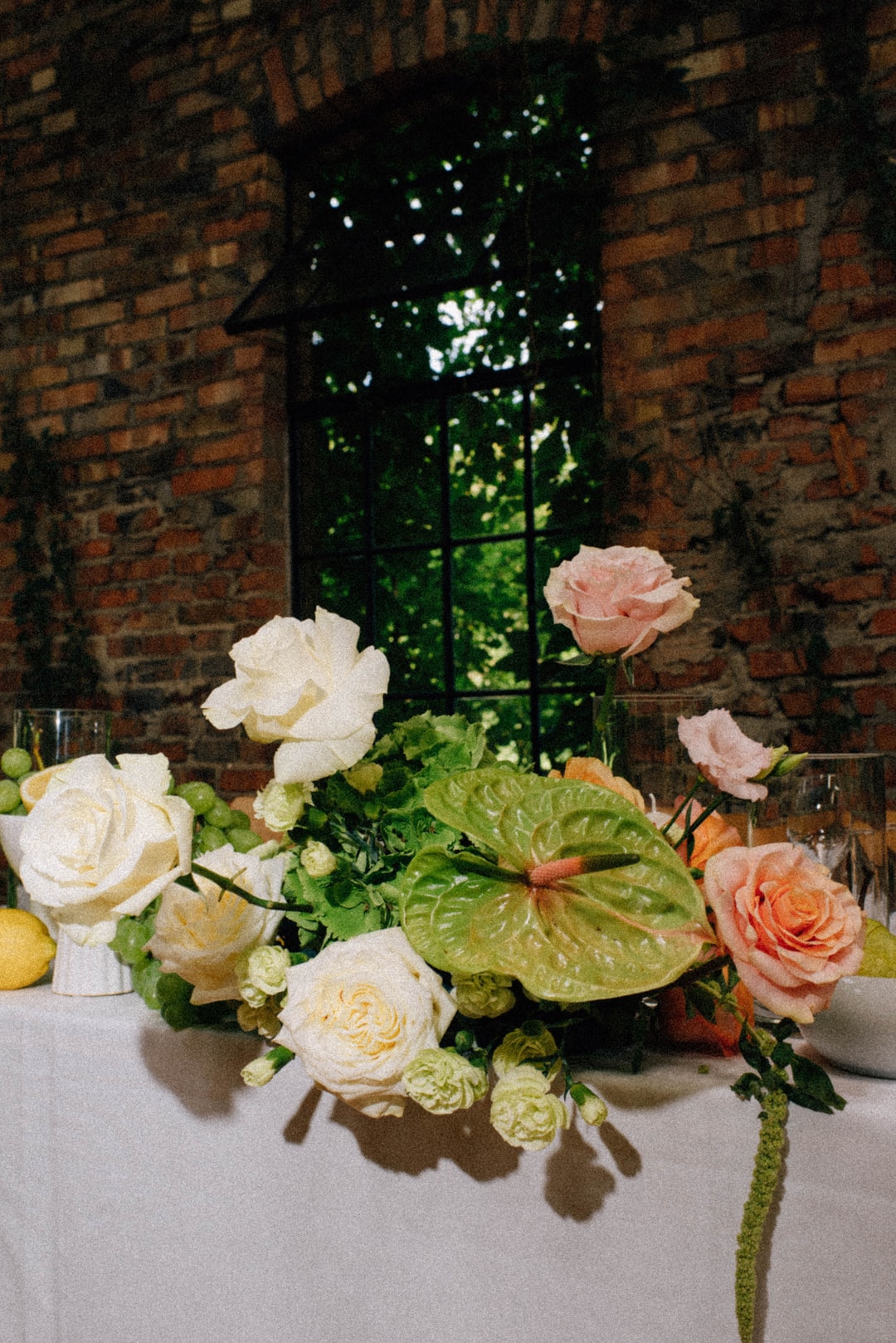 Kompozycja kwiatowa na stół z białymi i różowymi różami, zielenią oraz anturium, przy ceglanej ścianie z otworem okiennym.