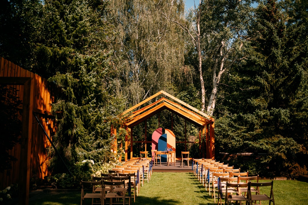 Drewniana altana z pomarańczowymi ramami wśród zieleni, drewniane krzesła ustawione jako dekoracje sali weselnej w ogrodzie.