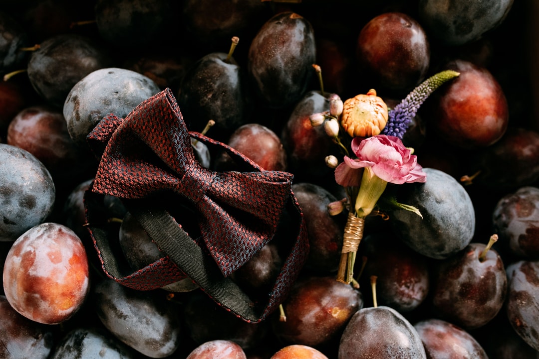 Kompozycja kwiatowa na stół z różowymi kwiatami, fioletowymi pąkami i bordo kokardą wśród owoców w ciepłej palecie.