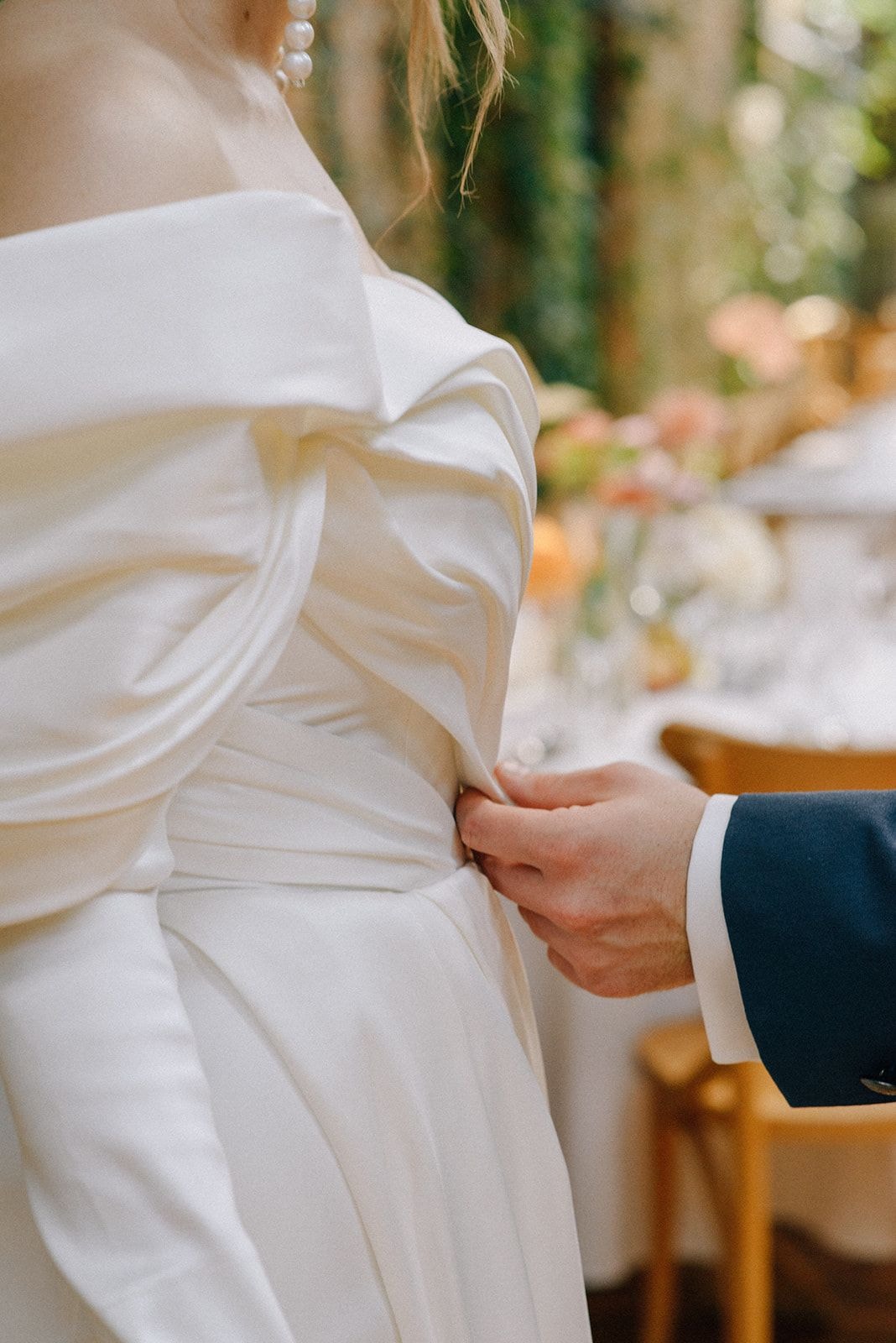 Panna młoda w białej sukni z draperią, pan młody w garniturze przytrzymuje suknię, dekoracja sali weselnej w tle.