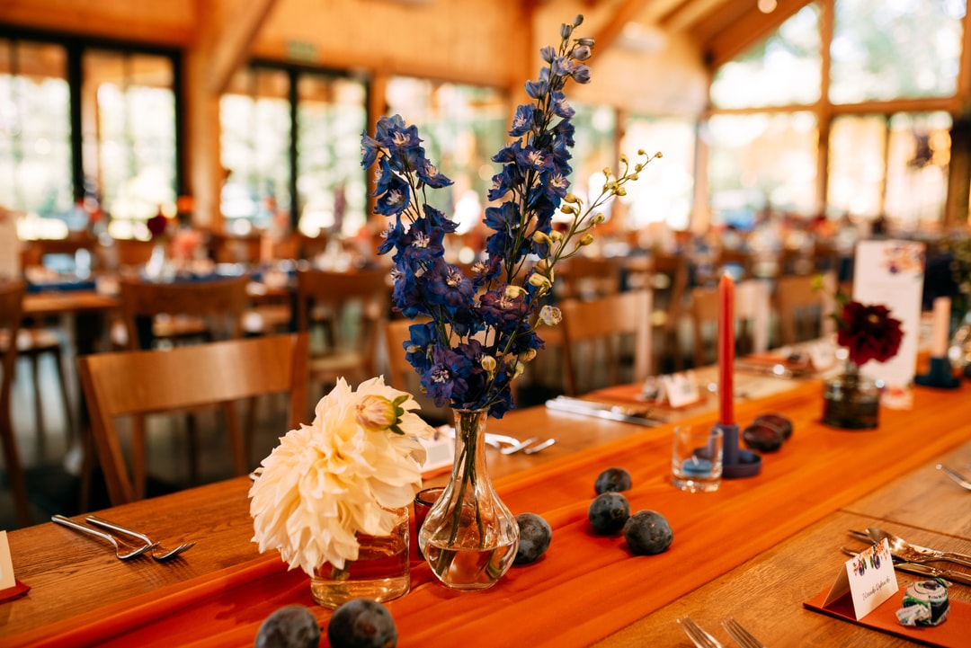 Aranżacja stołu weselnego z kompozycją kwiatową w szklanym wazonie, zawierającą kwiaty w odcieniach niebieskiego i białego na pomarańczowym bieżniku