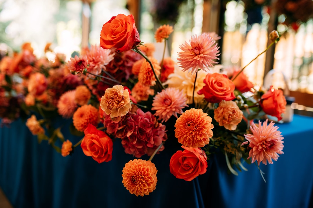 Kompozycja kwiatowa na stół z różami w odcieniach czerwieni i pomarańczu oraz dahlią, na tle niebieskiego obrusika