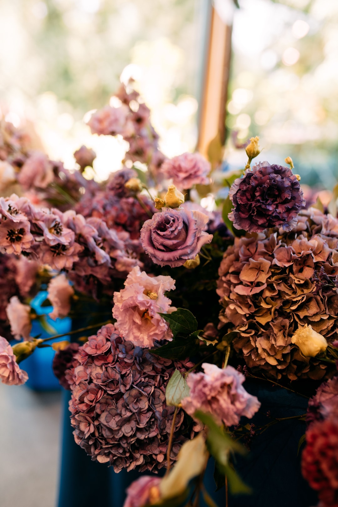 Zbliżenie kompozycji kwiatowej na stół w tonacji mauve i brązu, z różami i hortensją, w niebieskim wazonie