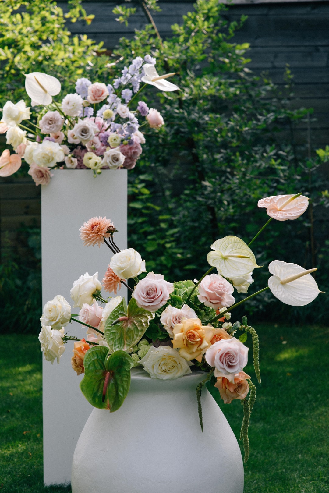 Dwupoziomowa dekoracja kwiatowa w białych ceramicznych wazonach. Górna kompozycja z różami, anturium i delphiniums w pastelowych tonach. Dolna czara z mieszanką kwiatów na stół w białej, różowej i pomarańczowej palecie.