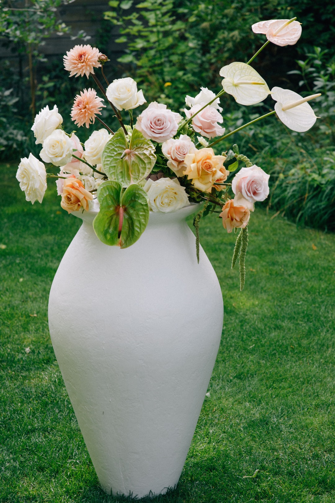 Kompozycja kwiatowa na stół w białym wazie z różami, dalią, anemonami i zielenią na tle ogrodu