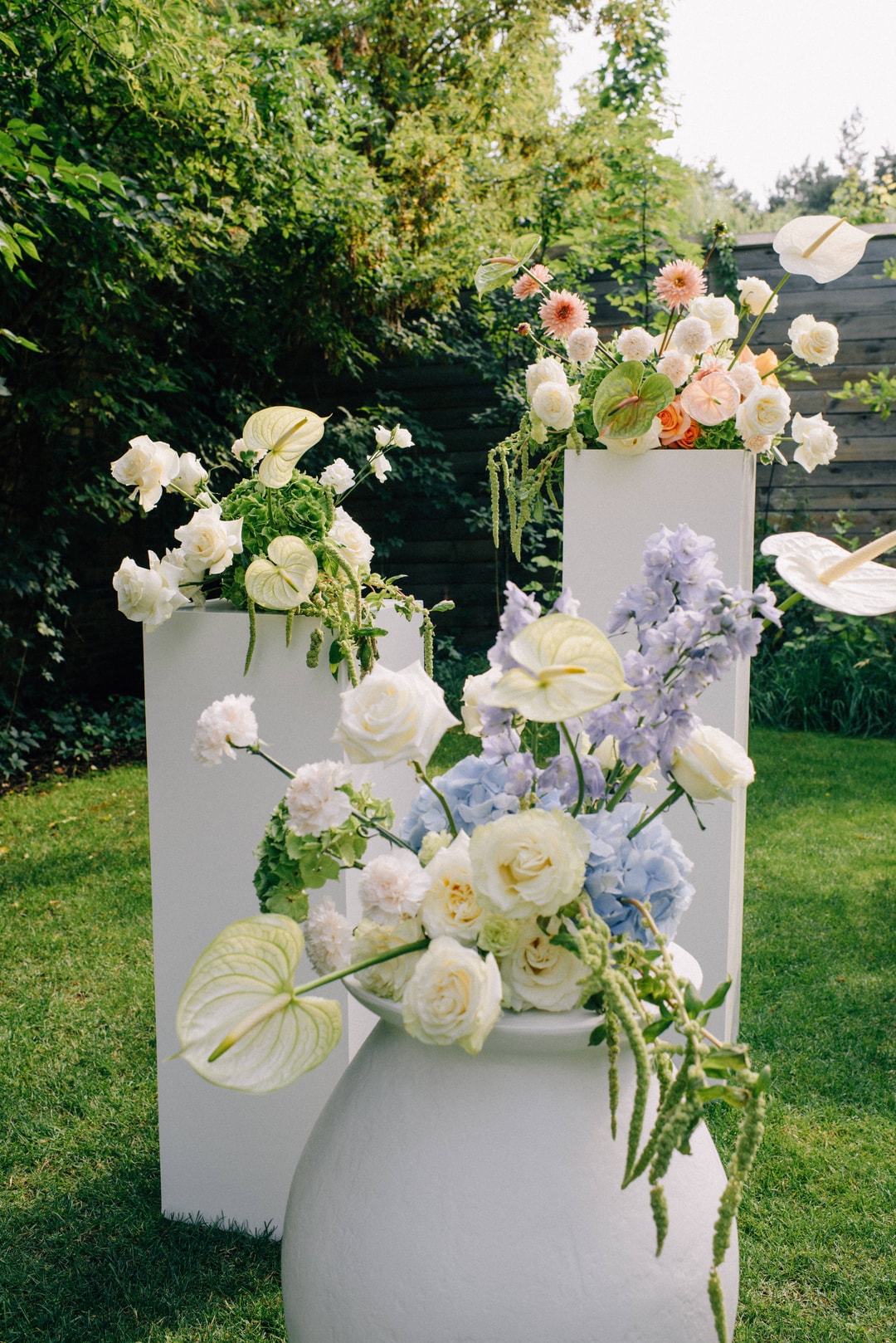 Trzystopniowa kompozycja kwiatowa na stół w białych i pastelowych tonach z różami, kwiatami polnymi i białymi anturium w ogrodzie.