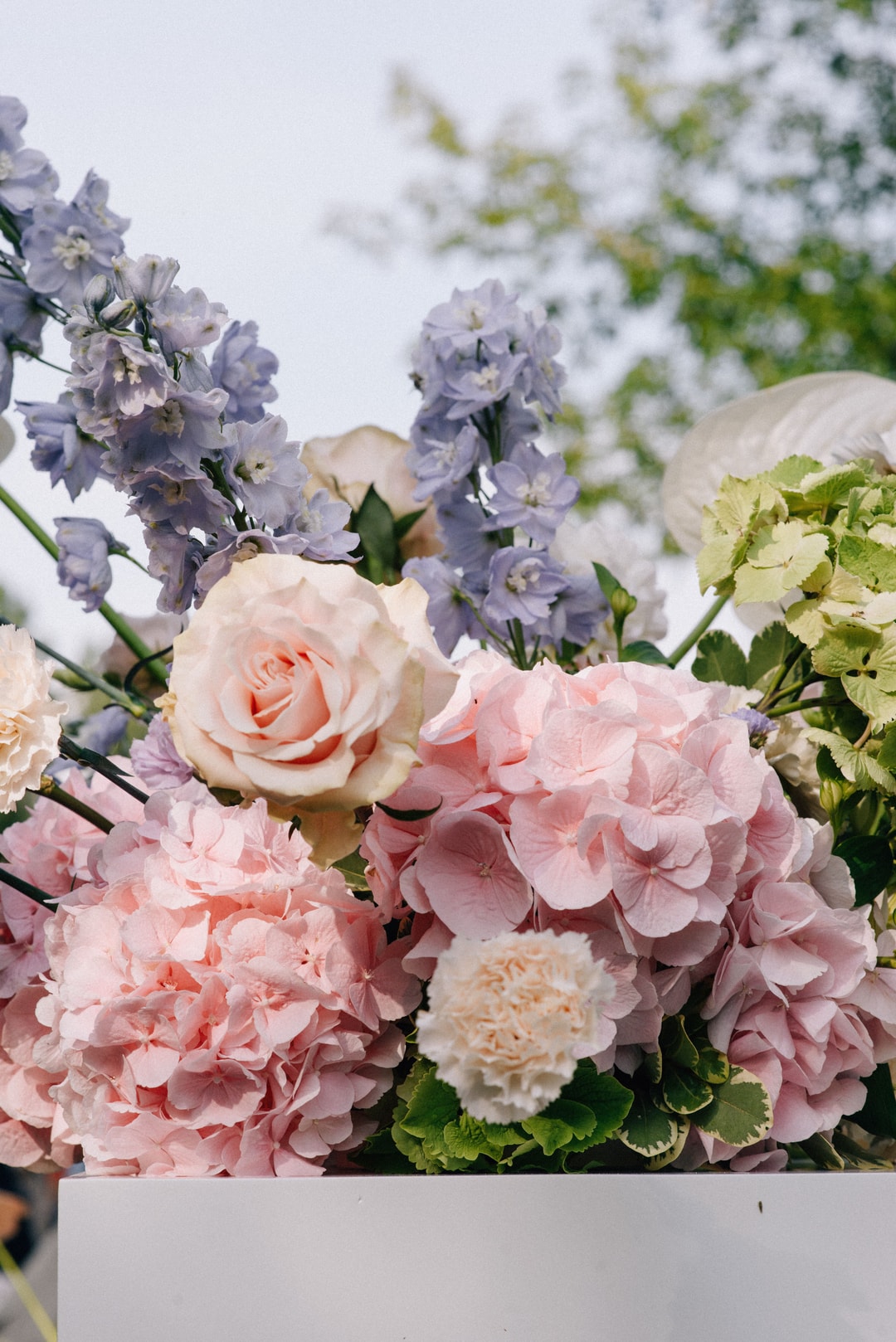 Kompozycja kwiatowa na stół z różowymi piwoniami, brzoskwiniowymi różami i lawendowymi kwiatami statice, liśćmi paproci i białą zielenią.
