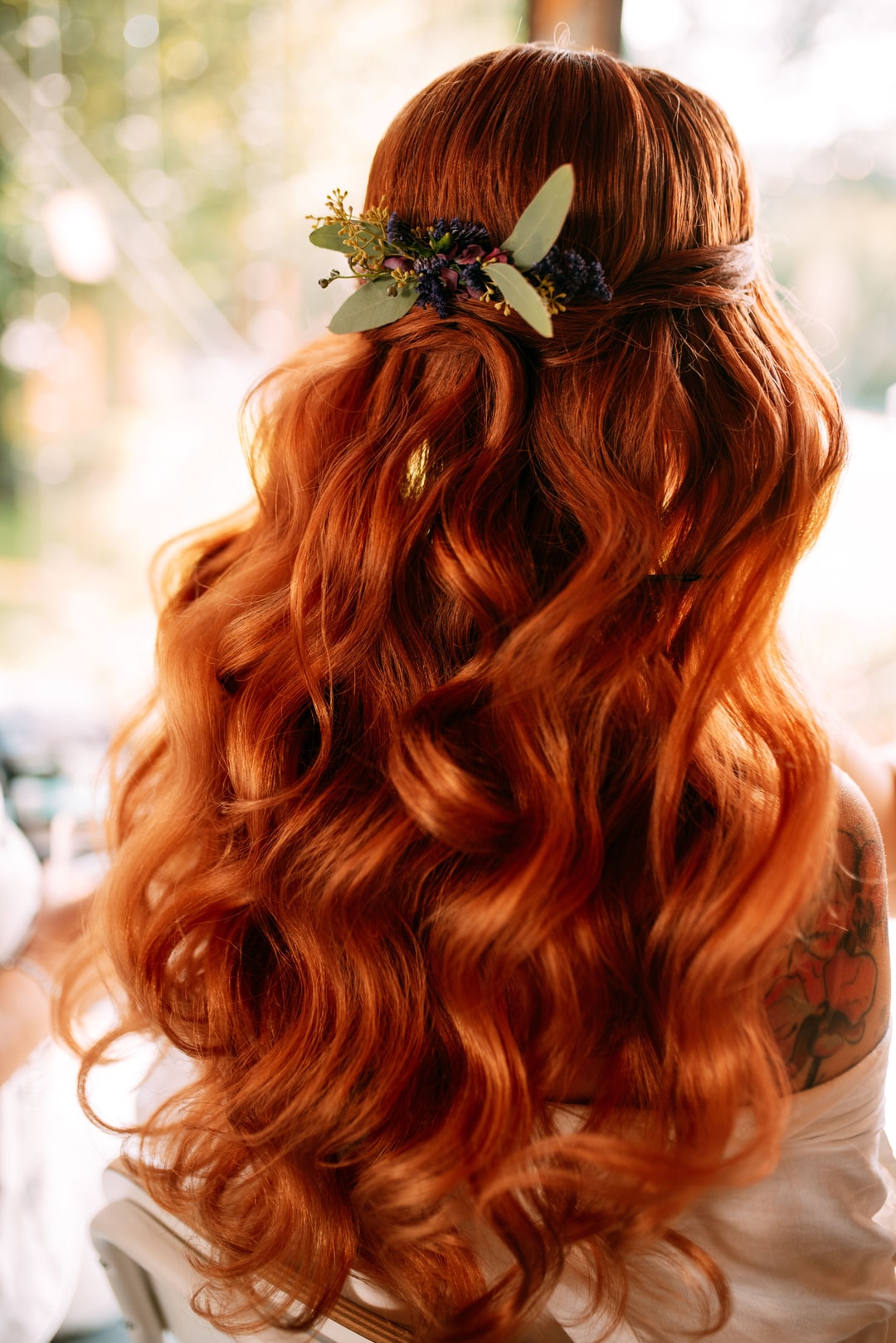 Długie rude włosy z falami i половinką upięcia, ozdobione zielonymi liśćmi i delikatnymi kwiatami w ciepłych tonach.