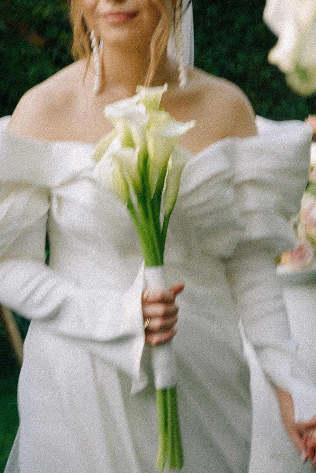 Panna młoda w białej sukni trzymająca bukiet ślubny z białymi kwiatami i zielenią, na tle zielonych dekoracji.