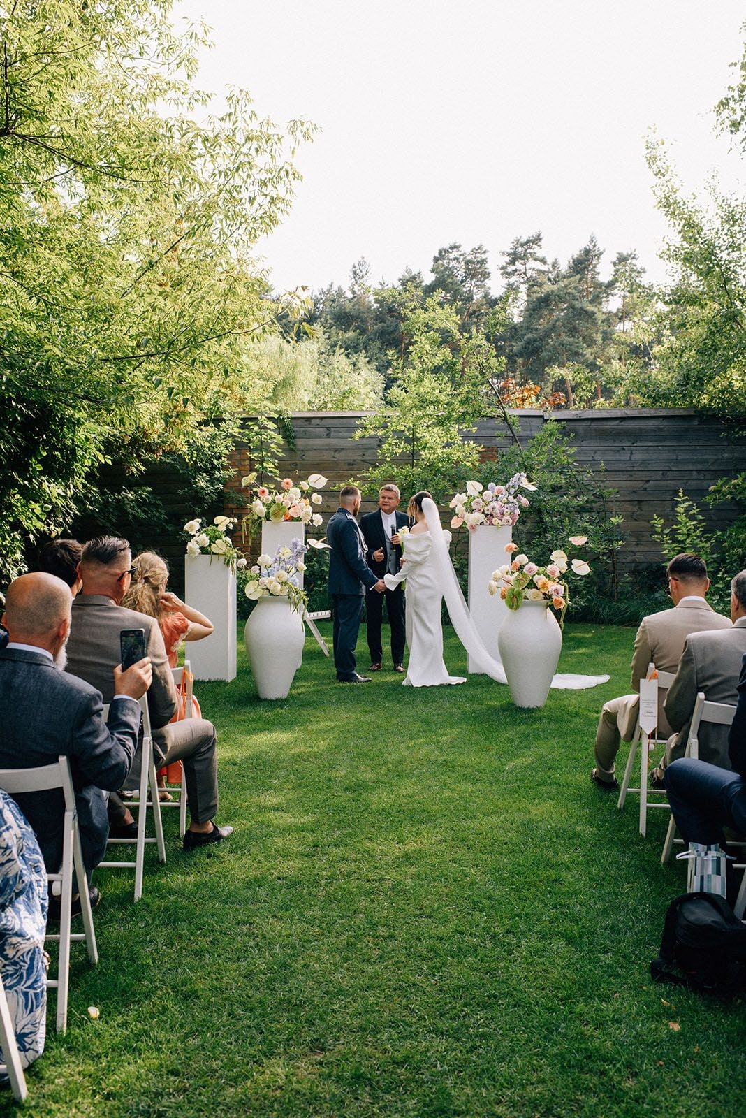 Ceremonia ślubna w ogrodzie z białymi wazonami pełnymi kwiatów, para młoda przy ołtarzu, goście siedzą po bokach alejki na trawie.