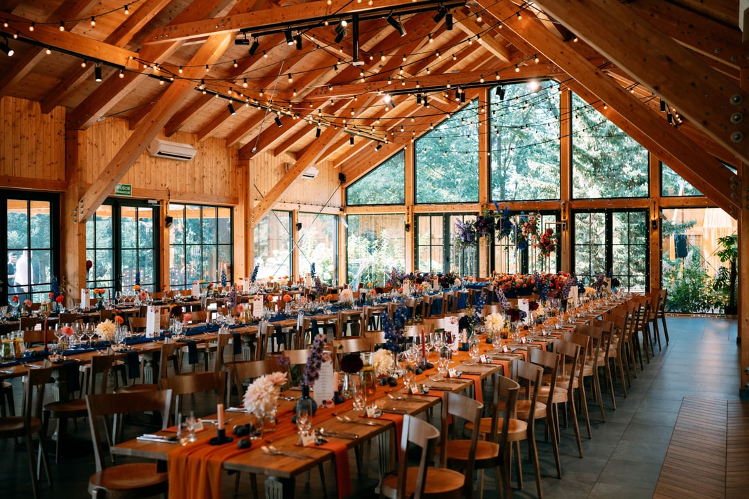 Drewniana sala weselna z dekoracjami stołów w pomarańczowych tonach, długie stoły ustawione w rzędach, goście przy stołach, duże okna z widokiem na las.