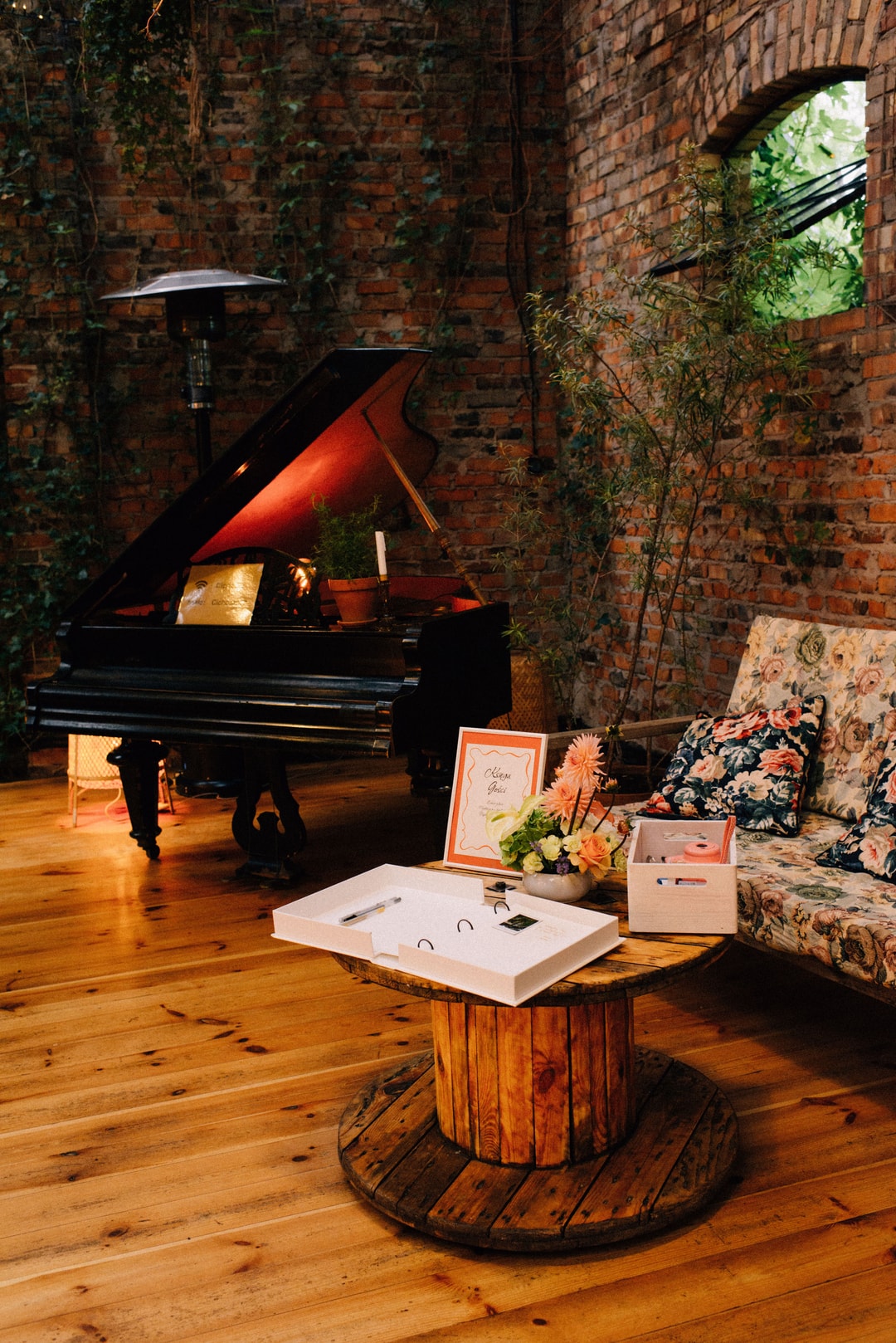 Wnętrze z ceglaną ścianą, czarny fortepian, drewniana podłoga, kompozycja kwiatowa na drewnianym stole, poduszki, dekoracje sali weselnej.