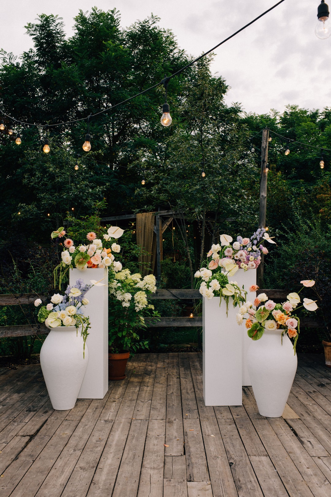 Cztery białe wazon-słupy z dekoracjami kwiatowymi w jasnej palecie na drewnianej podłodze, otoczone zielenią i światłami, tworząc aranżację sali weselnej