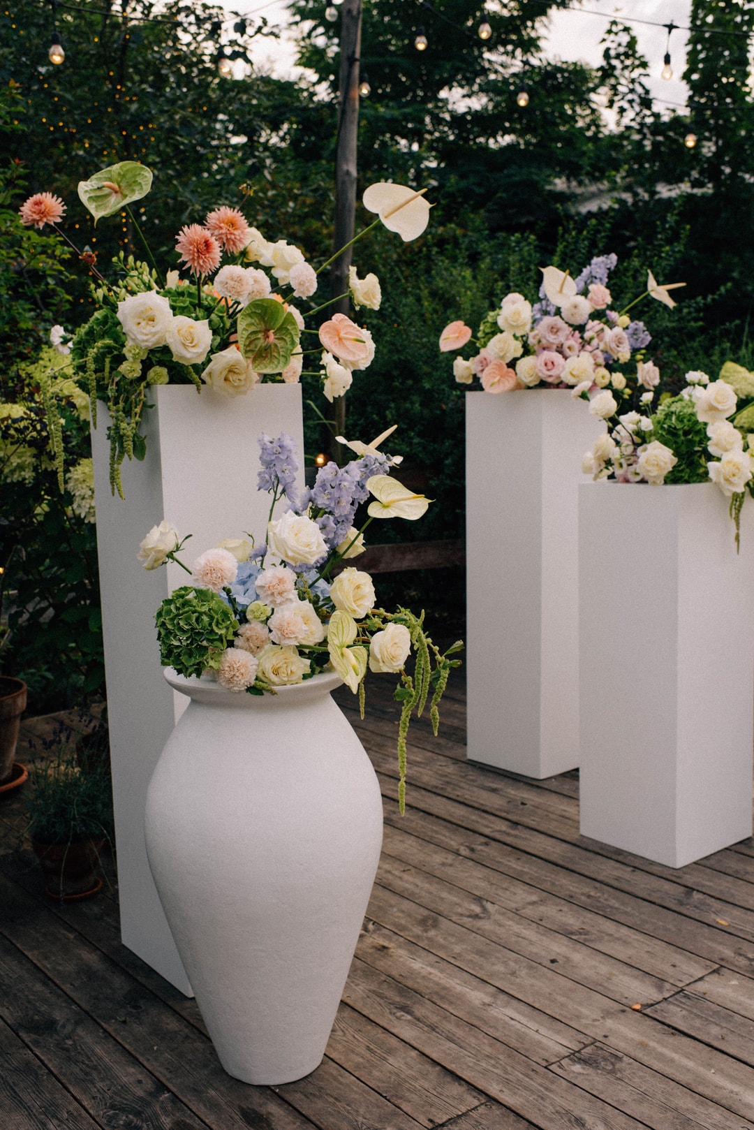 Trzy białe posągi z dekoracją kwiatową na drewnianej podłodze w ogrodzie. Kompozycje zawierają kwiaty w jasnej palecie z zielenią.