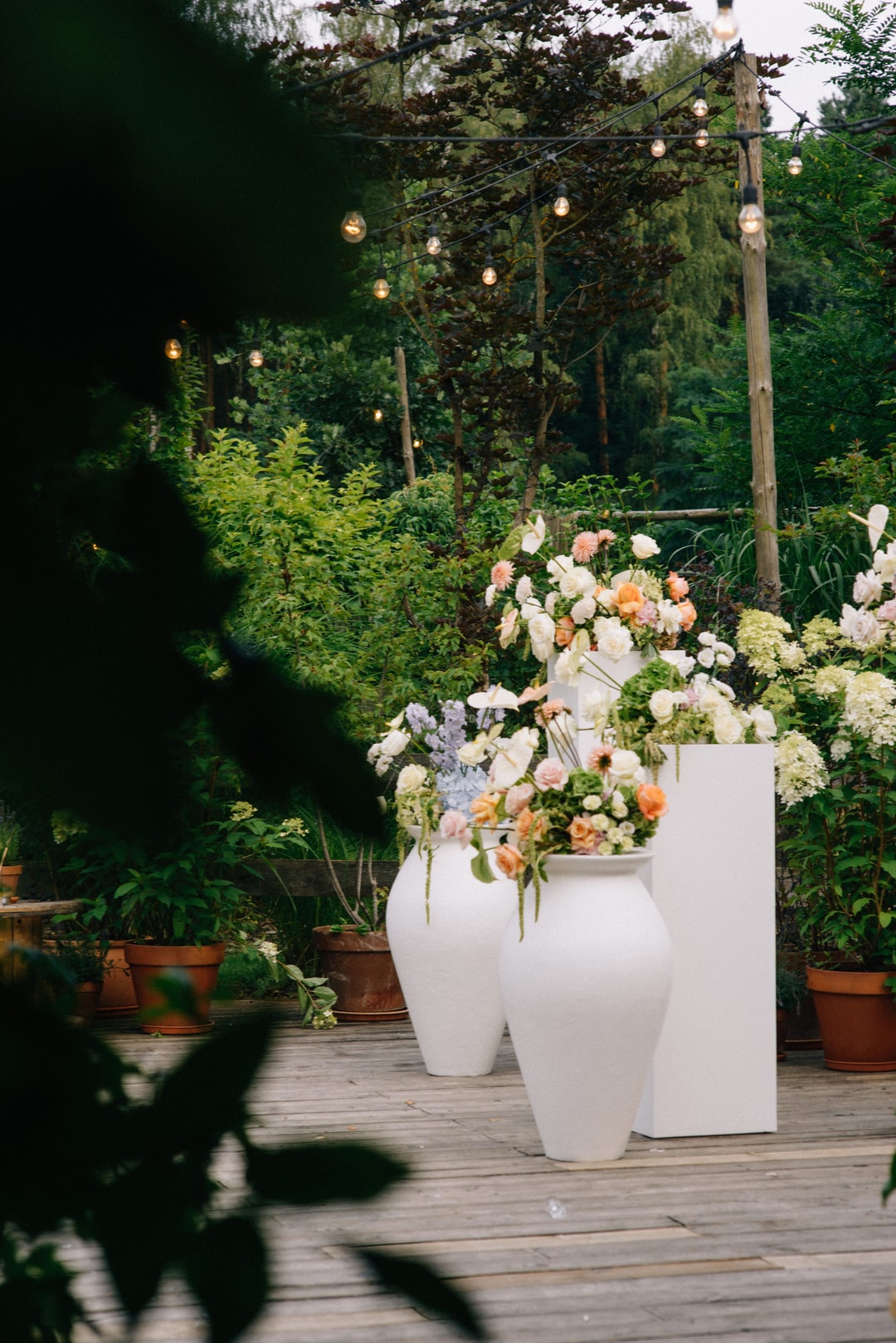 Białe pojemniki z kompozycją kwiatową na stół, zawierające kwiaty w jasnej palecie, zielone rośliny, drewniana podłoga, girlanda zieleni w tle