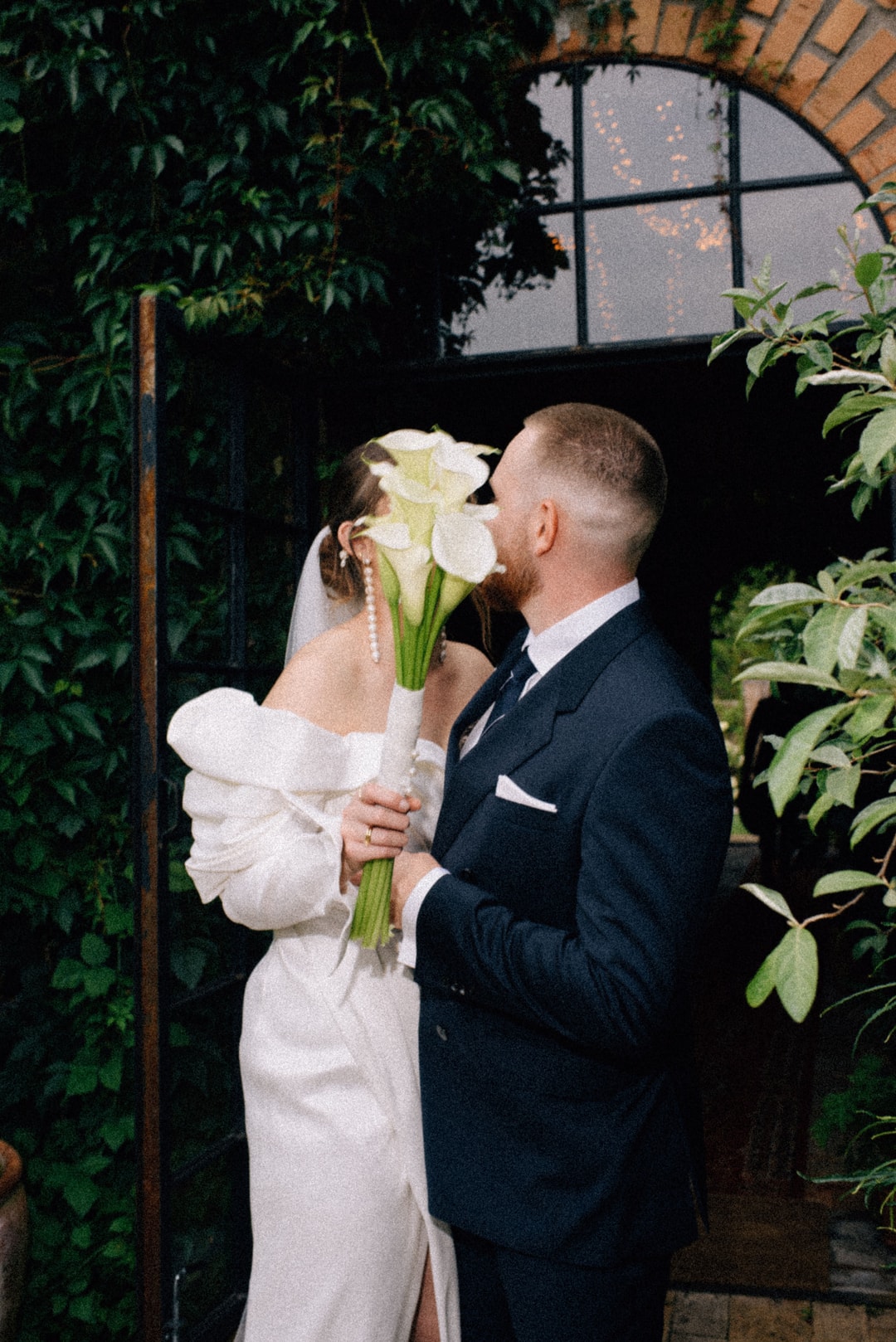 Para Młoda z bukietem ślubnym białych kalek, kobieta w białej sukni, mężczyzna w granatowym garniturze, zielone rośliny na tle.