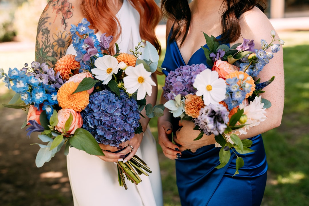 Dwie osoby trzymające bukiety ślubne z niebieskimi hortensją, białymi stokrotkami, pomarańczowymi dalią i fioletowymi kwiatami na tle ogrodu.