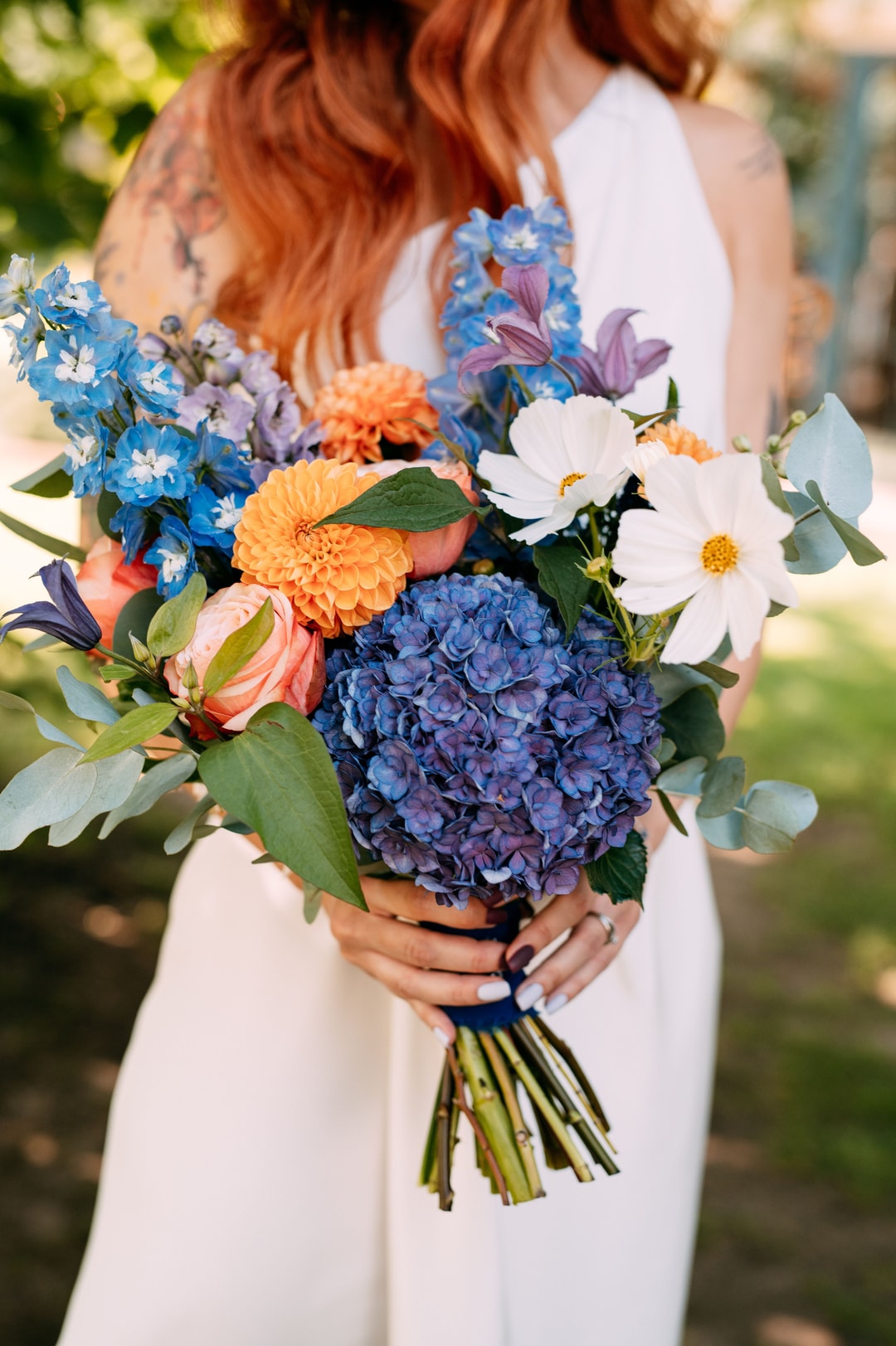 Bukiet ślubny z hortensją w odcieniach niebieskiego, kwiatami w jasnej palecie i zielenią, trzymany przez pannę młodą w białej sukni.