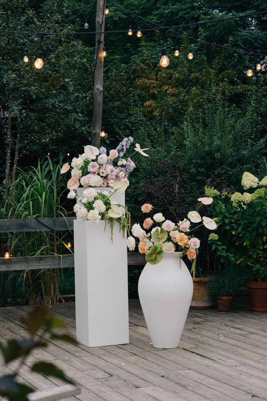 Dwie białe wazony z dekoracją kwiatową na stół – kwiaty w jasnej palecie na drewnianym tarasie z zielenią w tle