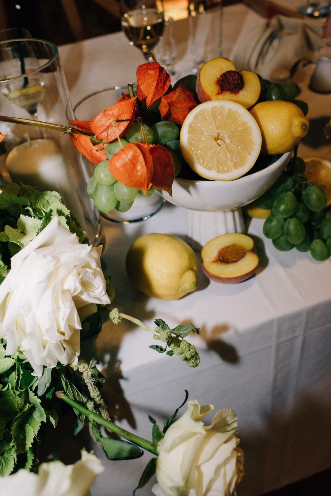 Dekoracja stołu z białymi kwiatami, cytrynami połówkami, brzoskwiniami i pomarańczowymi lampionami, zielonymi winogronami na jasnym obrusie.