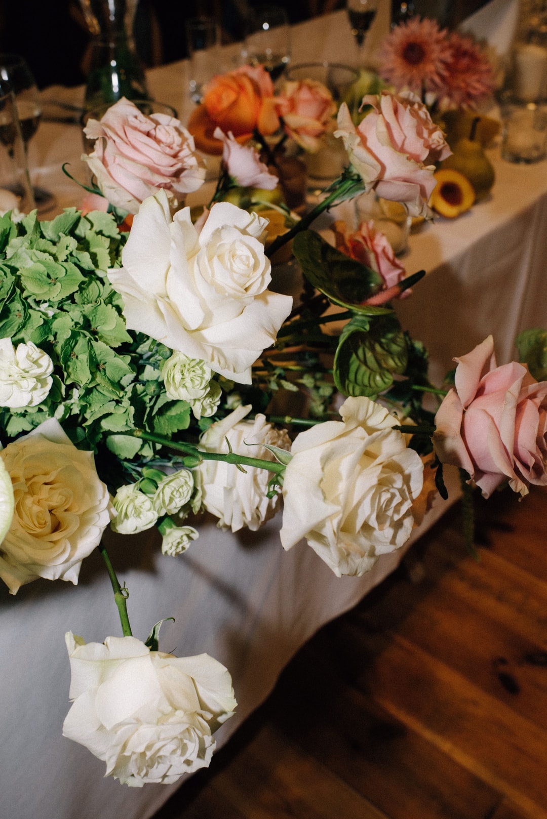 Kompozycja kwiatowa na stół z białymi różami, różowymi różami, żółtymi różami i zielenią dekoracyjną na szarej tkaninie.