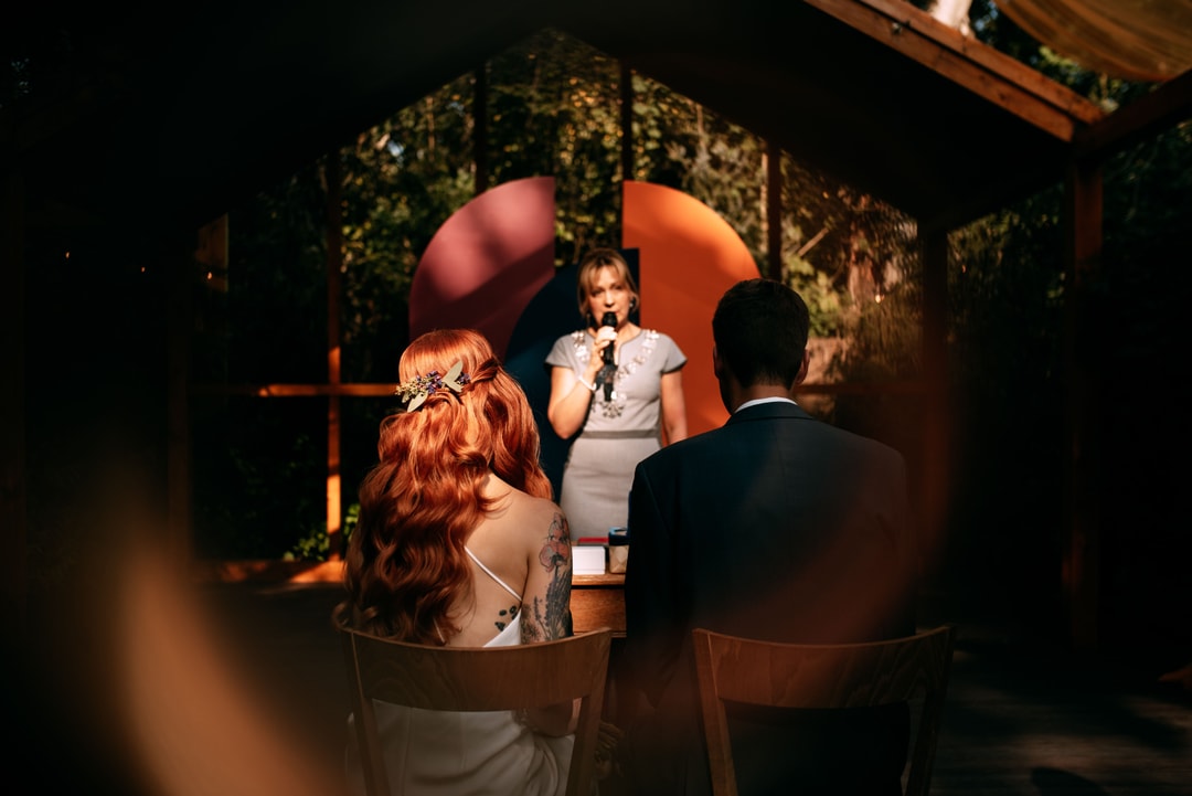 Kobieta w białej sukni mówi do mikrofonu przed parą gości, tło dekoracyjne z zielenią i jasnym łukiem kwiatowym.