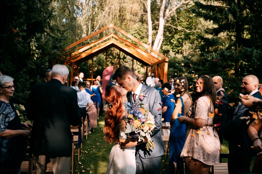 Para Młoda całuje się pod drewnianym łukiem kwiatowym w otoczeniu gości podczas ceremonii ślubnej w ogrodzie.