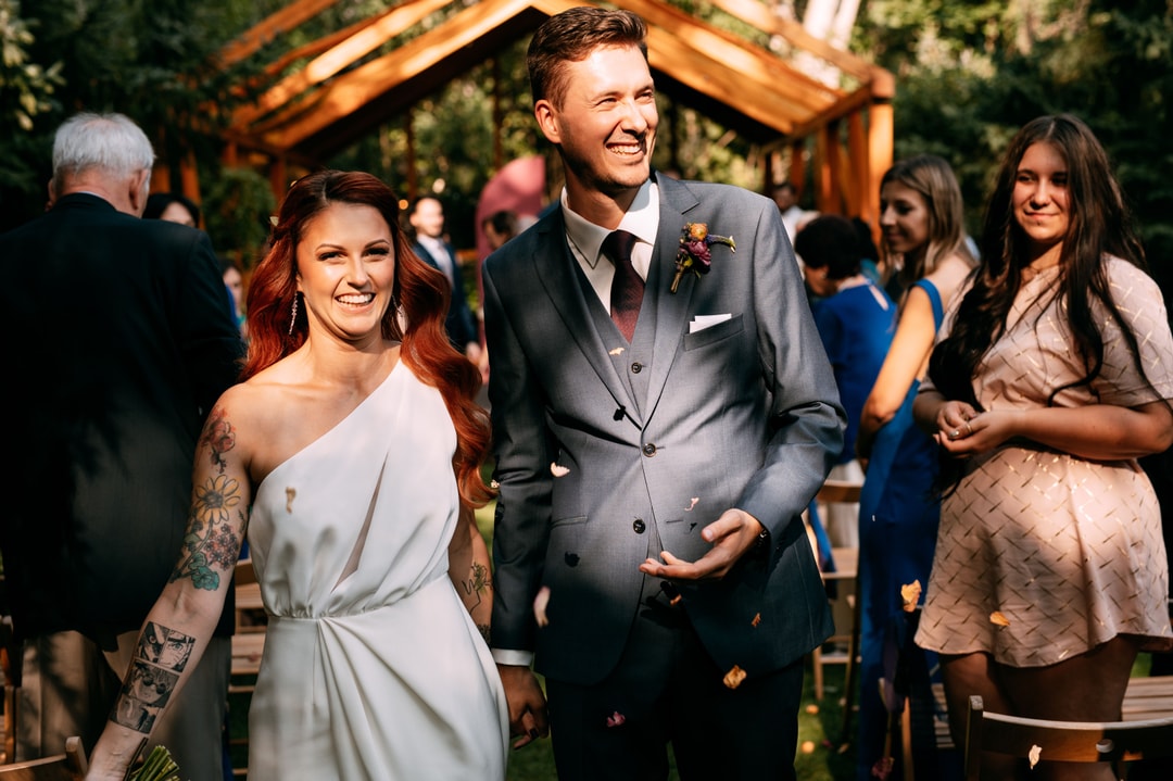 Para Młoda spaceruje alejką weselną pod drewnianym łukiem kwiatowym z zielenią, uśmiechająca się do siebie, goście obserwują ceremonię.