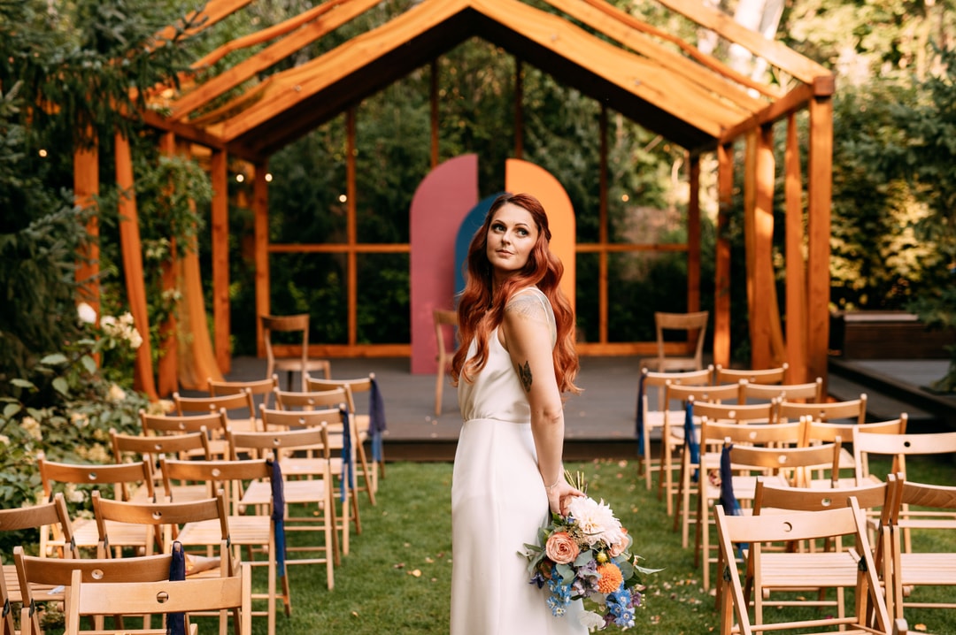 Panna młoda w białej sukni z bukietem kwiatów stoi na alejce weselnej między krzesłami, drewniany łuk kwiatowy w tle