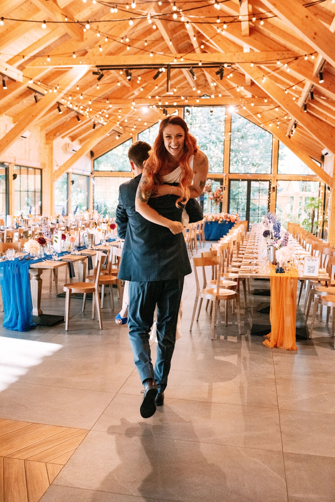 Mężczyzna w garniturze podnosi kobietę w czarnej sukni w drewnianej sali weselnej z dekoracjami kwiatowymi i oświetleniem.