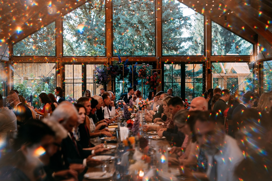 Dekoracje sali weselnej z długimi stołami, gośćmi w czarnych strojach, kwiatowymi kompozycjami, tło z dużych okien i zieleni.