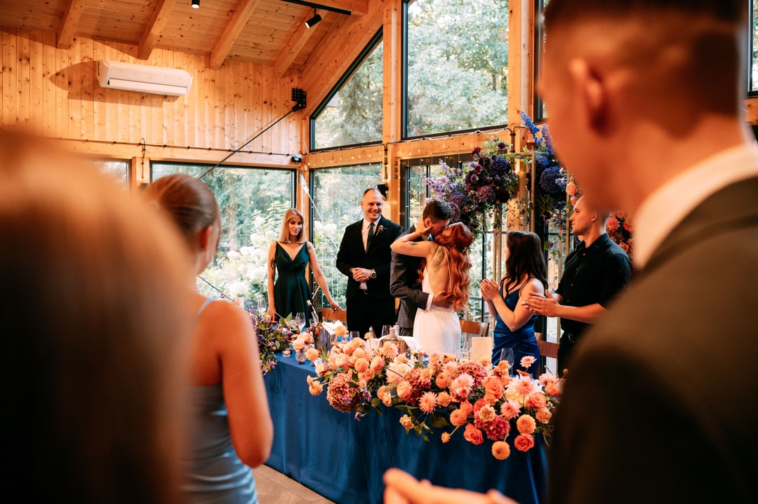 Dekoracje sali weselnej z kompozycją kwiatową na stole w jasnej palecie, drewniana sala z panoramicznymi oknami, goście i personel weselny