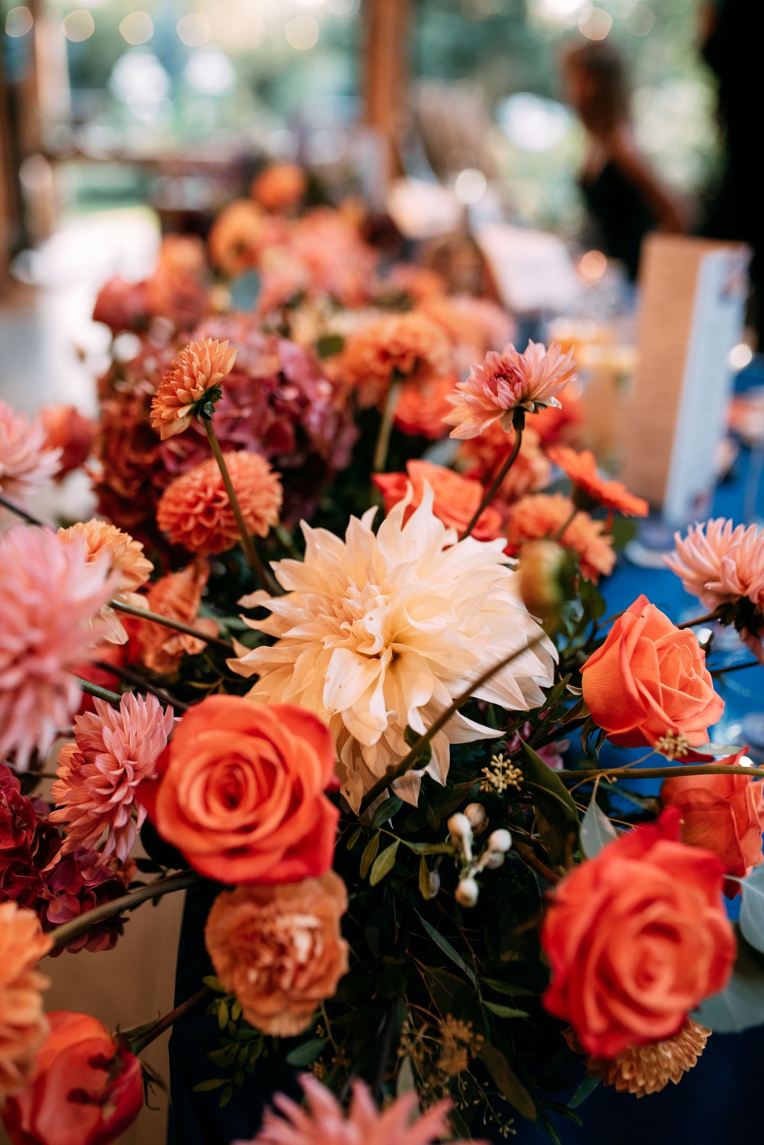 Kompozycja kwiatowa na stół z różami, daliami i kwiatami w odcieniach pomarańczy, czerwieni i szampana na tle dekorowanej sali.