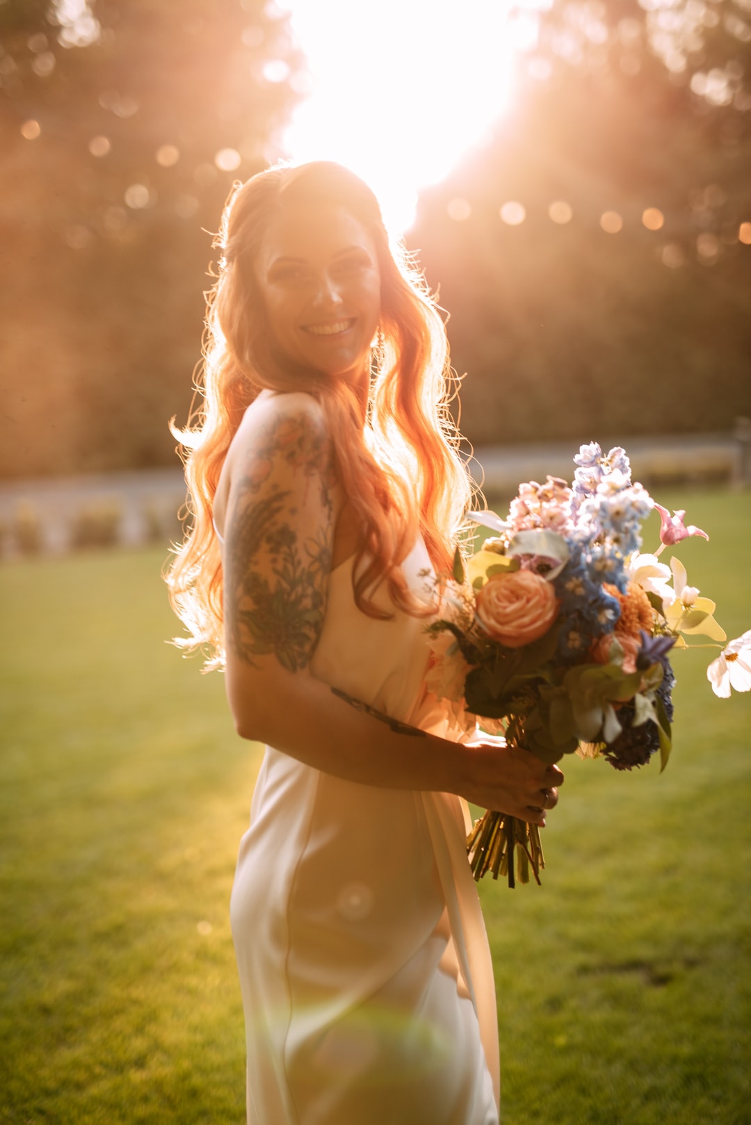 Kobieta w białej sukni trzymająca bukiet ślubny z kwiatami w jasnej i ciepłej palecie, na tle oświetlonego ogrodu