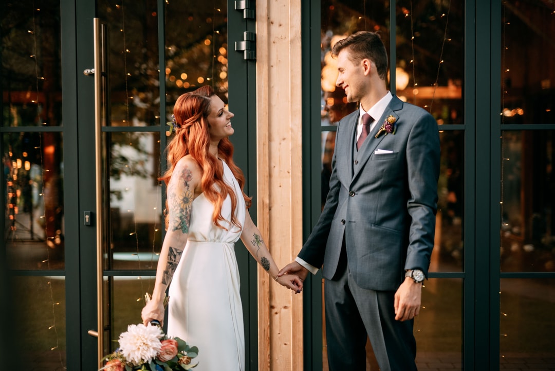 Para w strojach ślubnych trzymająca się za ręce przed wejściem, букет ślubny z jasnych kwiatów w dłoni panny młodej, wieczorowe światła w tle